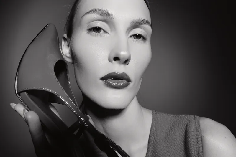 Close-up of a pale skinned white woman with slicked hair holding a high heel shoe near face in monochrome. Concept of fashion, elegance, and minimalism.