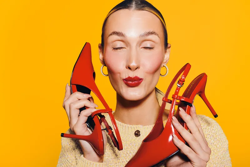 Young woman with closed eyes holds red high heel shoes near face on bright yellow background. Concept of fashion, style, footwear, and joyful emotions in studio lighting.