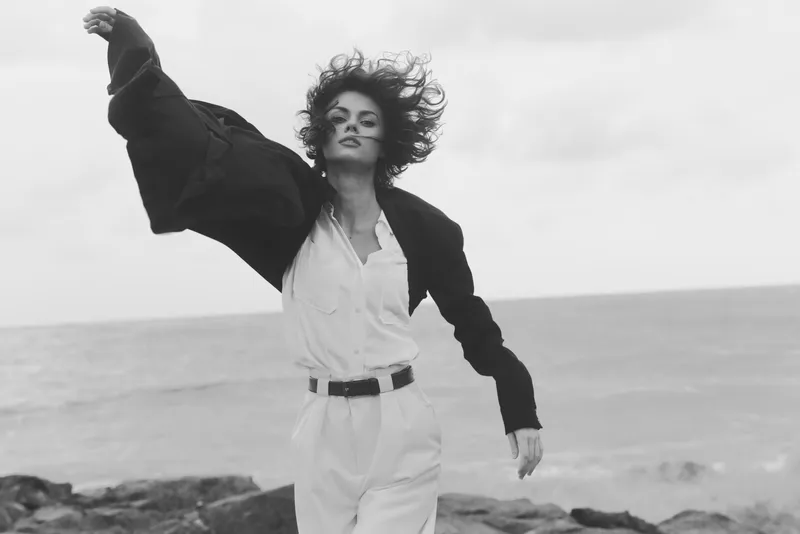 Woman on a windy beach wearing a white shirt and blazer, expressing freedom in motion as waves crash against rocks and the horizon frames a dynamic coastal mood.