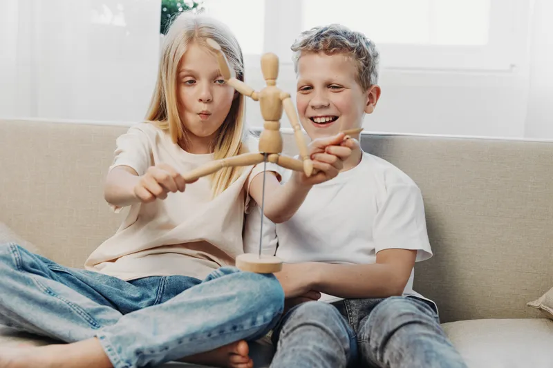 Two happy children sitting on a couch playing with a wooden artist mannequin, enjoying creativity and fun time in a bright living room environment.