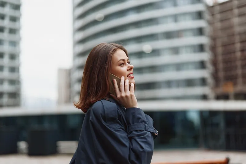 Young woman with long brown hair talks on smartphone outdoors in urban environment wearing dark jacket and smiling with positive expression.