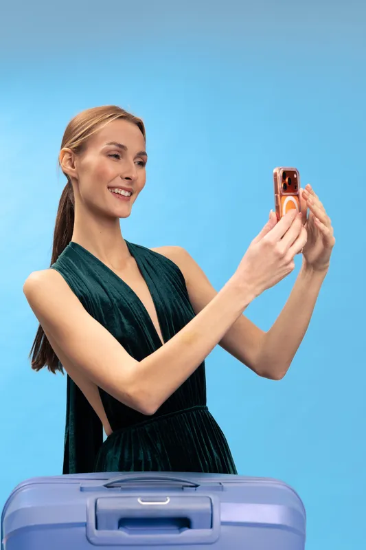 Young woman in elegant dress takes selfie with smartphone, smiling and standing behind suitcase on blue background. Concept of travel, vacation, and holiday preparations.