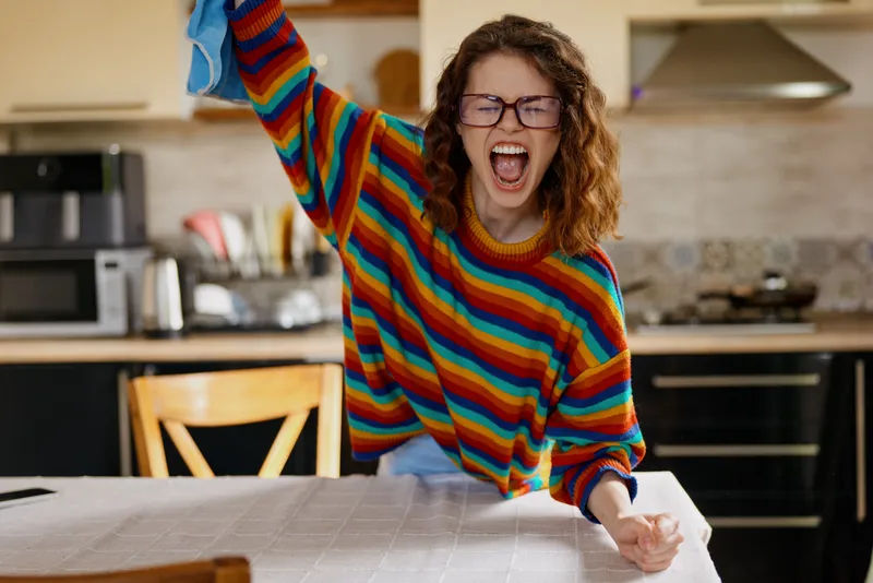 A cheerful woman in a colorful striped sweater stands at a kitchen table, fists raised in excitement. A bright, welcoming room conveys upbeat home energy and a playful, joyful mood.