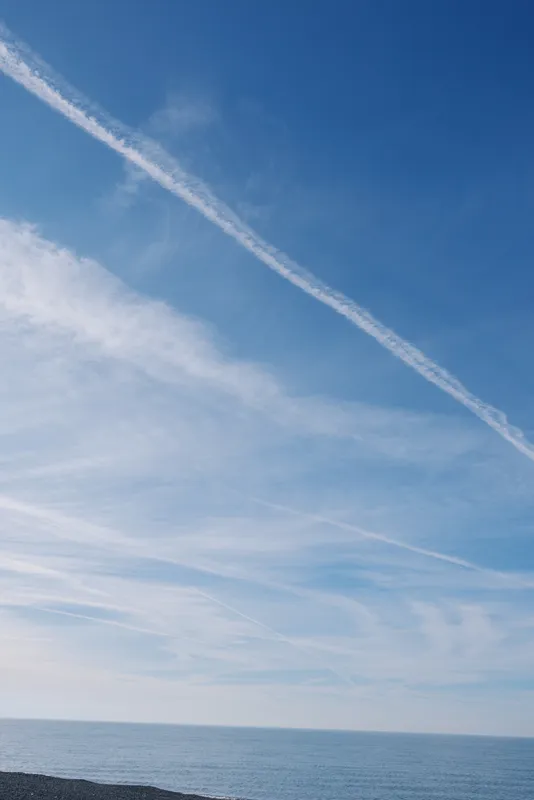 Sky and sea stretch under a blue expanse with white contrails crossing the calm horizon, clouds drifting over a tranquil coastal scene in a minimalist, peaceful outdoor setting