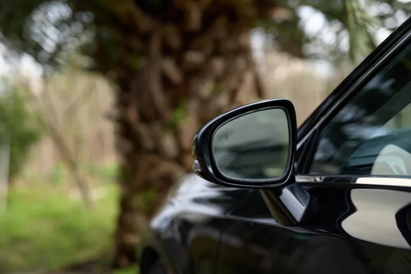 Close up of a black car side mirror outdoors, with a soft green background, capturing a calm reflective moment as a gentle breeze rustles nearby leaves along a shaded garden path in warm afternoon