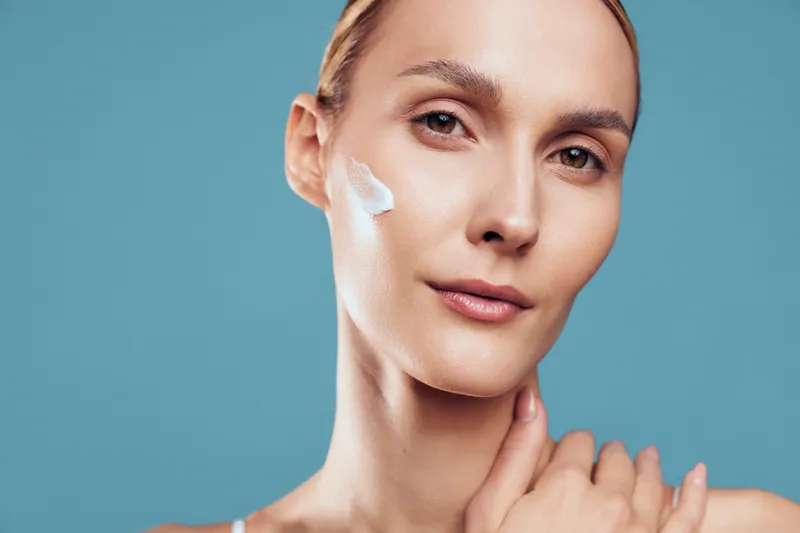 Young woman with clear skin applying white face cream on cheek, close-up portrait on blue background. Concept of skincare, beauty routine, and natural wellness. Soft lighting highlights healthy skin.