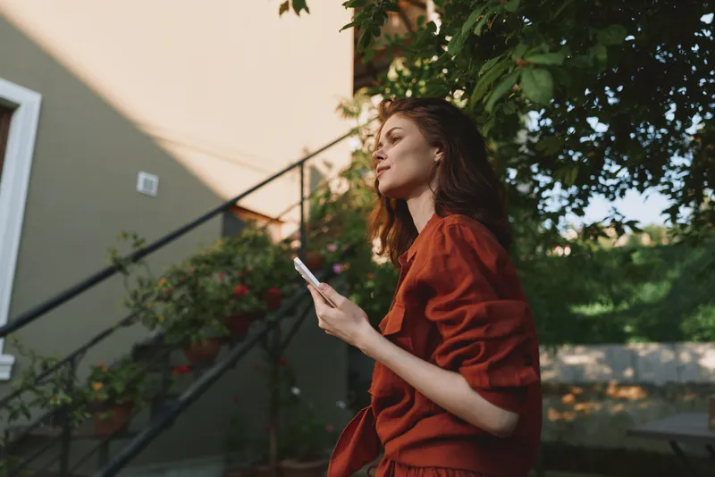 Woman lifestyle scene outdoors in warm light, wearing red outfit, holds a smartphone, relaxed pose, candid moment, calm garden backdrop, natural mood, contemporary style