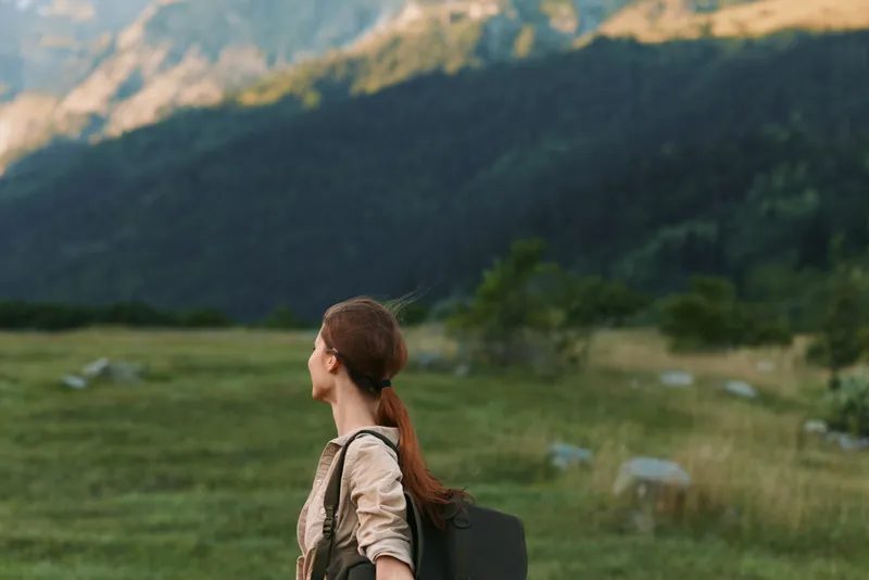 Woman traveler with backpack gazes toward distant mountains in a grassy meadow, embracing outdoor lifestyle and serene nature scene.