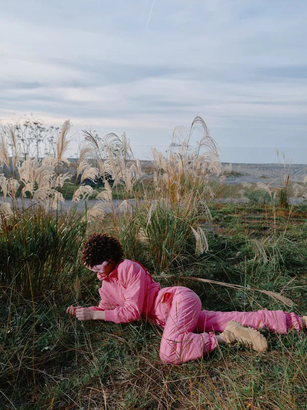 Person lying on grass in an open field wearing pink clothing, outdoor scene, relaxed pose, natural surroundings under a cloudy sky, peaceful countryside mood and calm atmosphere.
