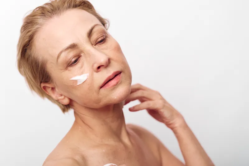 Mature woman with short blond hair applies white face cream on cheek. Soft natural skincare concept with focus on anti aging, wellness, and healthy skin care routine against plain background.