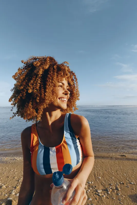 Young woman with curly hair smiles happily while standing on a sandy beach near calm water holding a water bottle under clear blue sky during a sunny day.