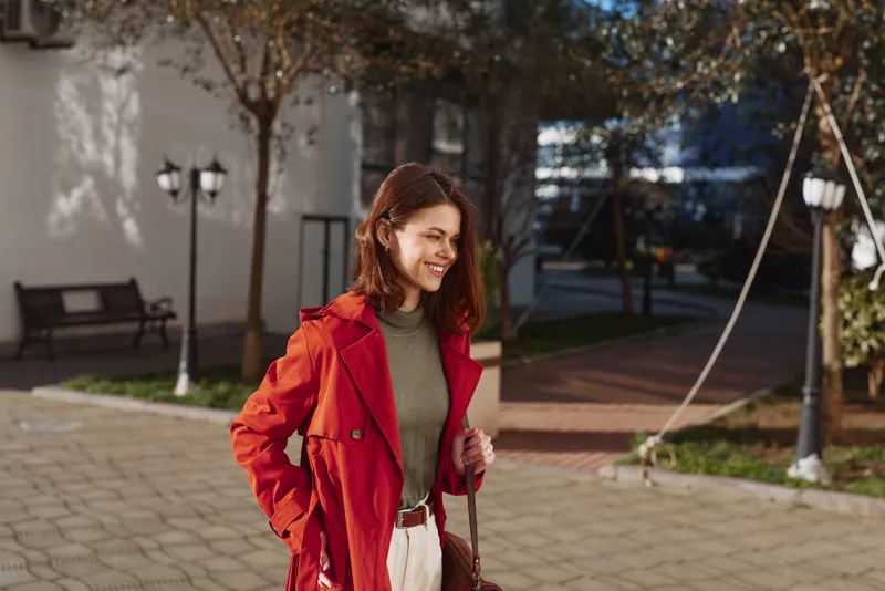 Woman in a red coat and green sweater walks outdoors in an urban park, smiling, casual fashion, candid street portrait with benches and trees in the background.