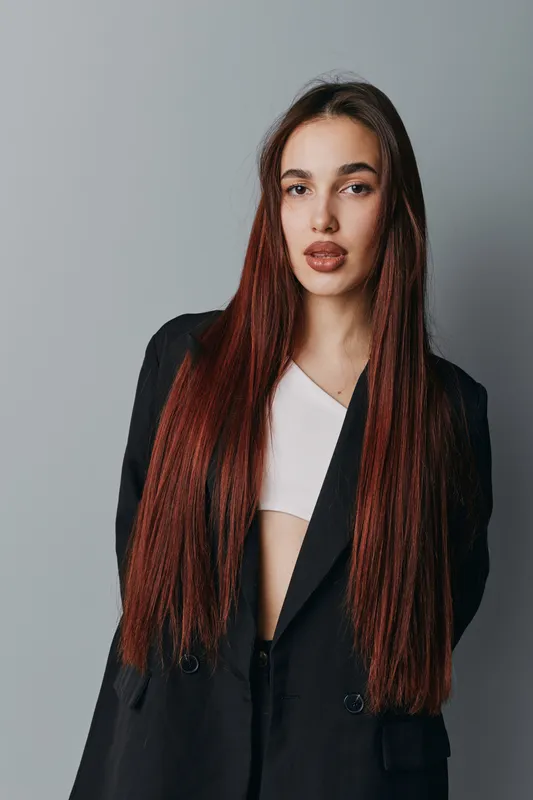 Young woman with long straight hair wearing black blazer and white top standing against gray background looking at camera with serious expression and confident posture.