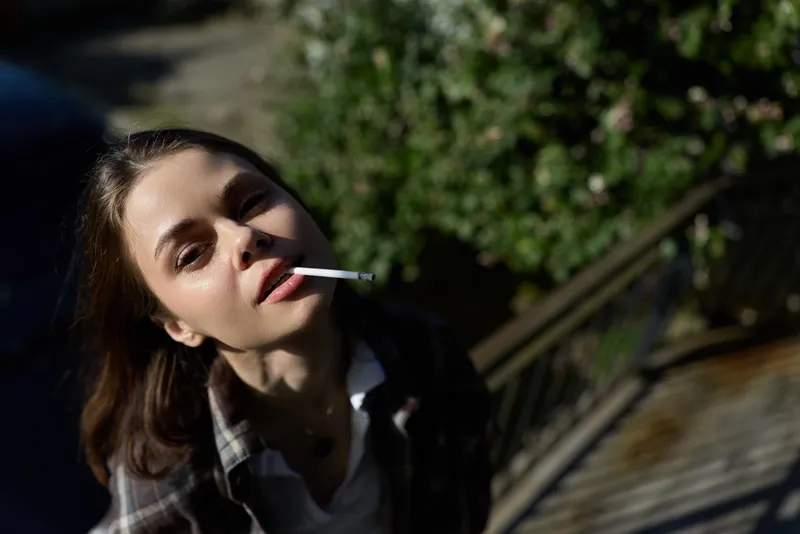 Young woman outdoors on sunlit stairs, cigarette in mouth, casual plaid shirt, relaxed gaze and informal mood