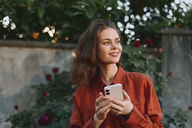 Relaxed young woman in an orange blouse smiles while holding a smartphone outdoors, surrounded by greenery and blooming flowers in a natural sunlit moment