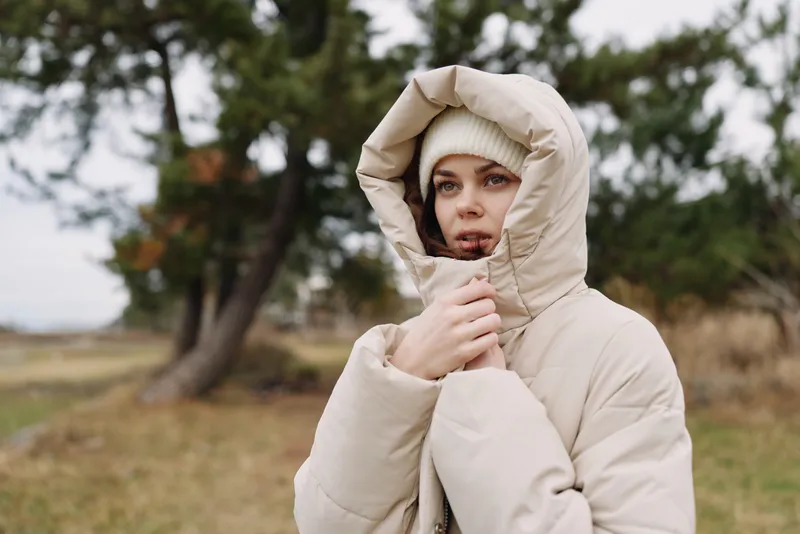 Outdoor portrait of a hooded woman in a beige puffer jacket in a chilly autumn field, calm expression under soft light. background trees for mood.