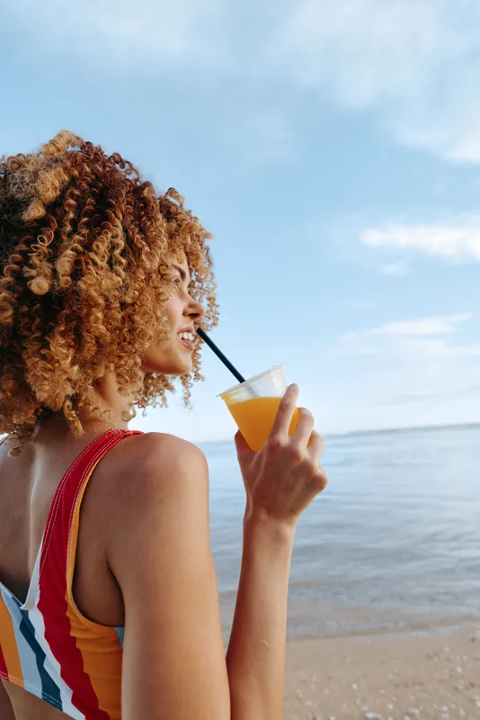 A young woman with curly hair enjoys a fresh orange drink by the beach on a sunny day. She wears a colorful swimsuit and looks relaxed while gazing at the sea.