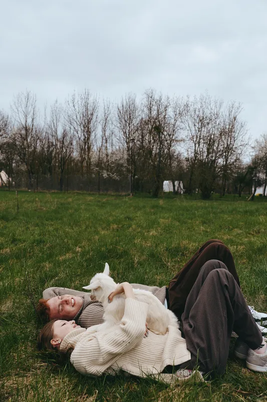 Goat kid rests beside a woman in a cozy sweater, lying on grass in a field, a relaxed lifestyle outdoor scene, a gentle intimate moment in nature with calm countryside vibes and warm, soft mood