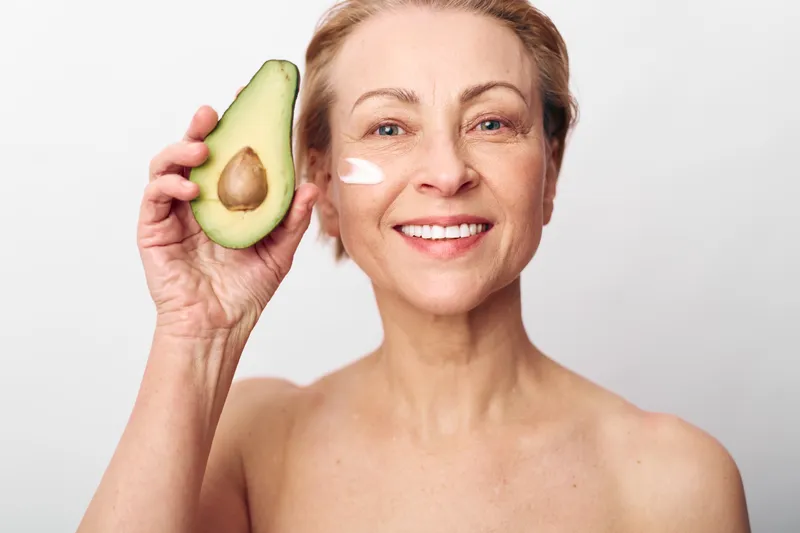 Smiling middle aged woman with natural skin holds half an avocado near her face while applying white cream on cheek. Concept of organic skincare, healthy lifestyle, and natural beauty care.