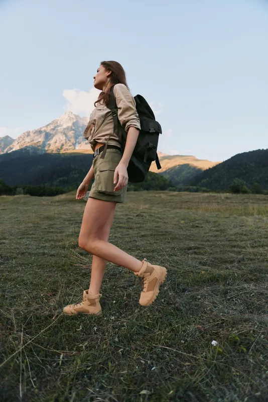 A young woman with a backpack strides across a grassy meadow toward distant mountains, exuding focus and determination as light washes the landscape and peaks glow softly.