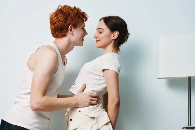 Young couple smiling and standing close facing each other in casual clothes at home with neutral wall and lamp on background. Intimate moment and connection between two people.