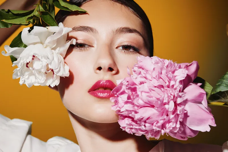 Close-up portrait of a fashionable young woman with glossy red lips and glowing skin, holding vibrant pink and white flowers against a warm orange background. Beauty and creative fashion concept.