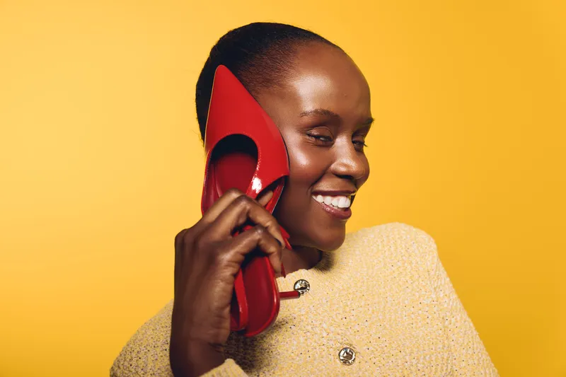 Smiling African woman with dark skin holds red high heel shoe near face on yellow background. Concept of fashion, beauty, happiness, and stylish accessories in studio lighting.