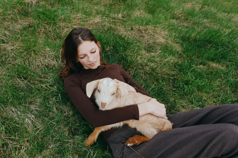 Joyful woman outdoors, resting on grass while holding a gentle goat kid, warm light, peaceful moment, close connection, casual attire, natural colors, relaxed mood, and gentle breeze