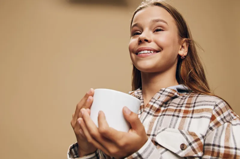 Woman smiling and holding a cup with both hands wearing a plaid shirt in cozy atmosphere. Portrait with happy expression and casual style on neutral background.