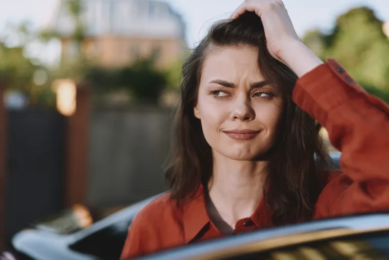 Young woman with brown hair sits in a parked car, touching her hair with a thoughtful smile, wearing a rust red shirt and looking off to the side on a sunny day