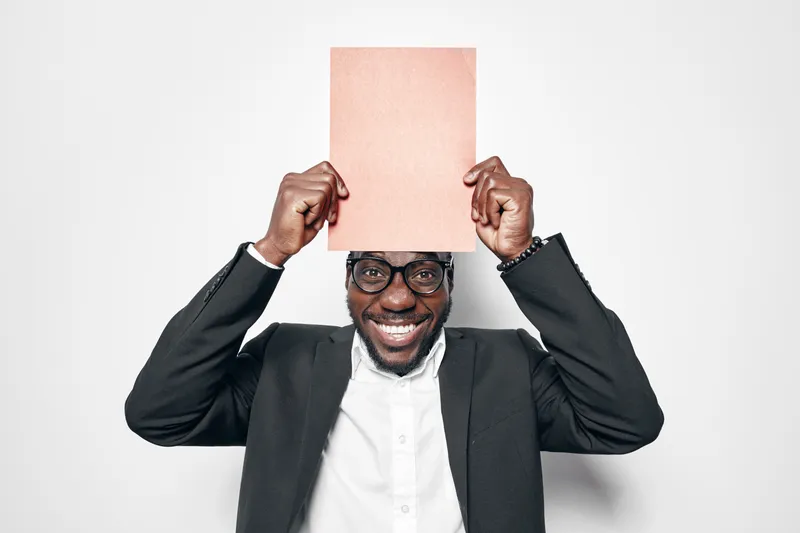 African man with dark skin wearing glasses and black suit holding blank paper above head smiling on white background. Portrait, happy, confident, casual and studio shot.