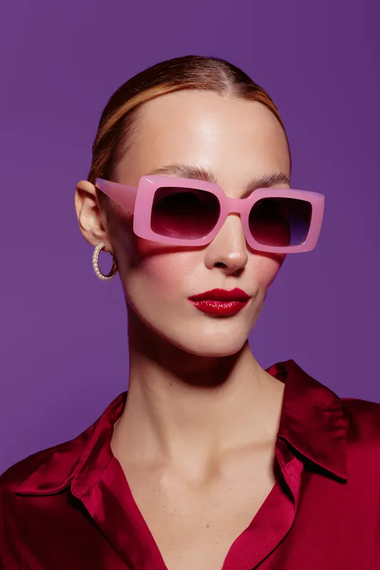 Stylish woman with pink sunglasses and red lipstick poses against purple background. Fashion portrait with elegant hairstyle and hoop earrings. Concept of glamour and modern style.