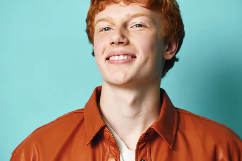 Young man with red hair and fair skin wearing brown leather jacket smiling confidently against blue background. Portrait, expression and casual fashion style in studio lighting.