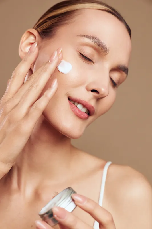 A young woman applies moisturizer from a small jar with her eyes closed and a subtle smile, bathed in warm soft lighting, conveying a calm skincare routine and self care moment.