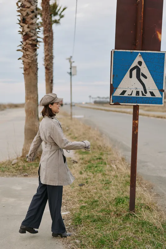Woman in stylish coat and hat walks on sidewalk near pedestrian crossing sign on quiet road with palm trees. Casual urban fashion concept with autumn or winter outdoor mood.