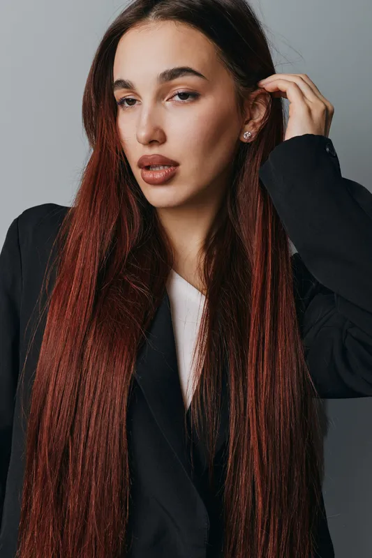 Woman with long straight red hair wearing black blazer and white top posing with hand near ear on gray background. Beauty portrait with natural makeup and confident expression in studio.