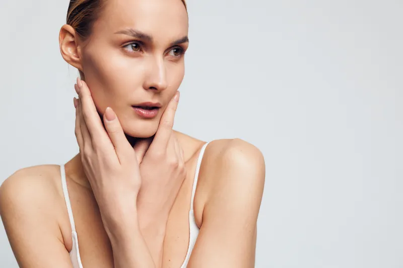 Young woman with clear skin touches her face gently while looking thoughtfully to the side against a plain light background in a white tank top.