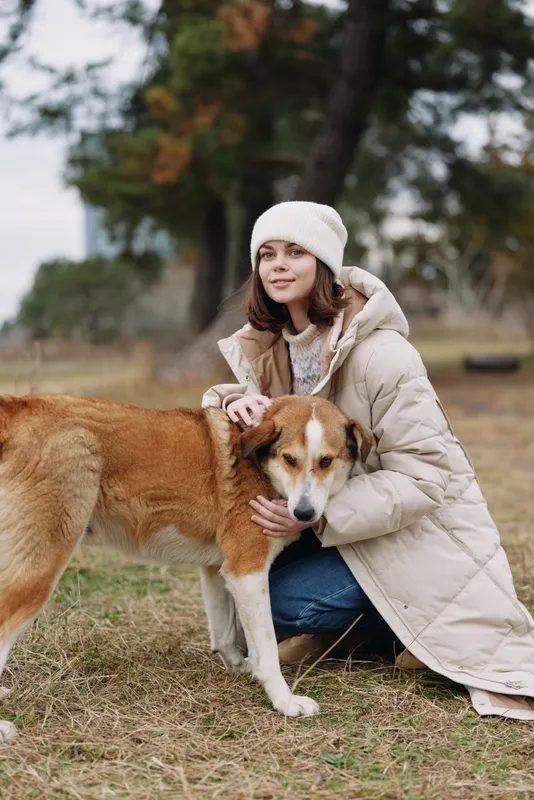 Woman kneels beside a dog in an open park, wearing a warm coat and beanie, offering gentle companionship in a calm autumn setting with soft natural light and a tranquil backdrop of trees.
