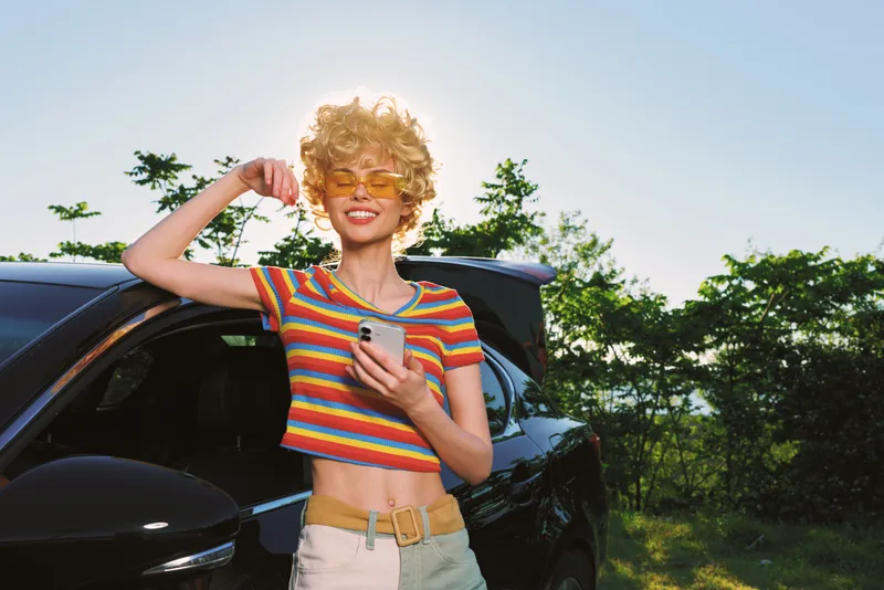 A stylish woman in a striped retro top stands by a dark car in bright outdoor sun, holding a smartphone. Concept for fashion, youth, and outdoor lifestyle.