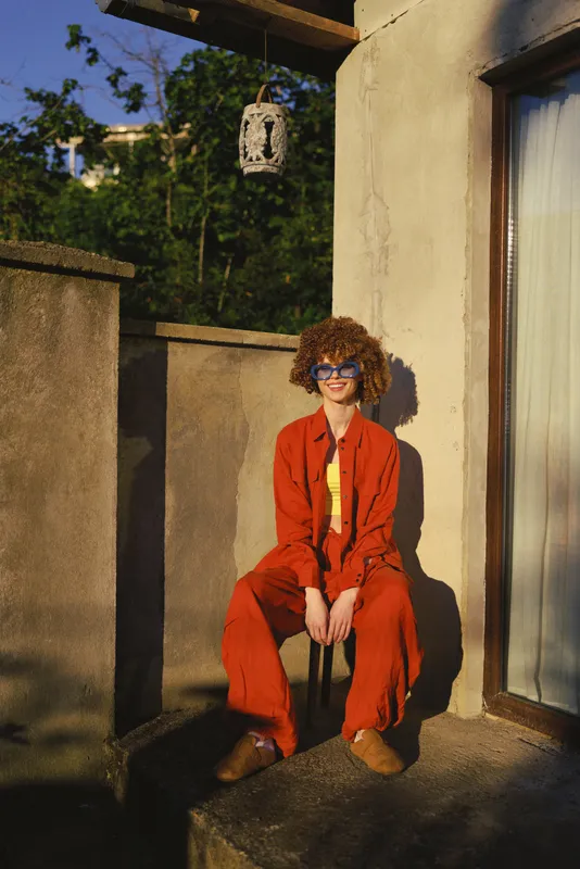 Cheerful woman outdoors, wearing a red suit and yellow top, sitting on a terrace, sunlit moment, casual fashion pose, lifestyle vibe