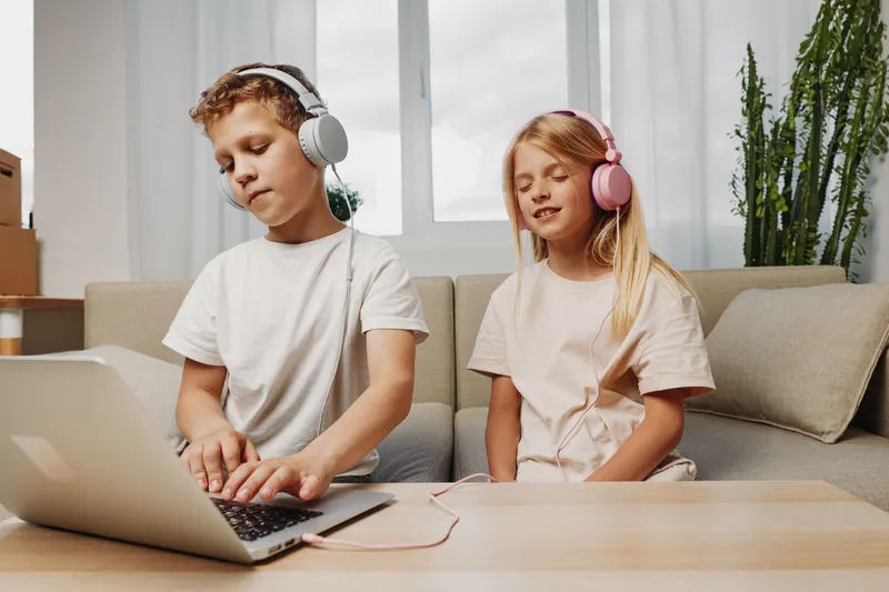 Two kids with headphones enjoying music and laptop on a cozy sofa, casual clothes, modern home interior with plants and natural daylight.