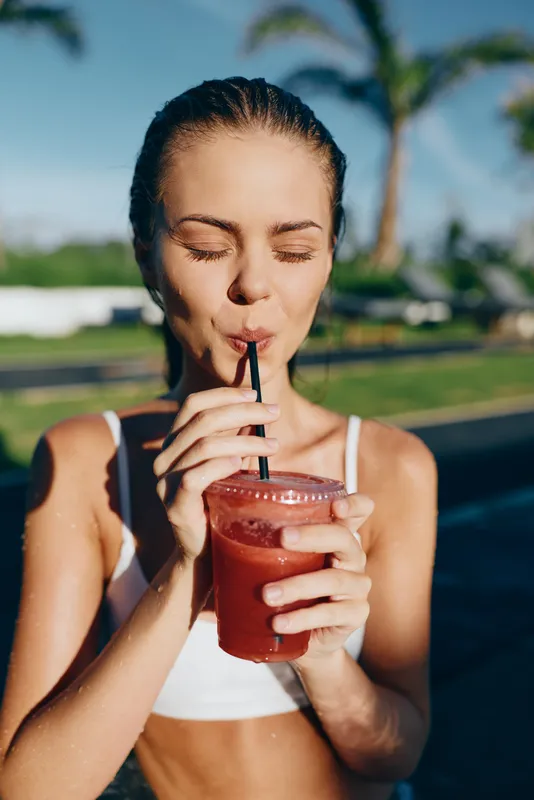 Cheerful woman outdoors sipping a fruity smoothie, eyes closed, sunlit moment, relaxed posture, tropical backdrop, vibrant colors, healthy lifestyle vibe