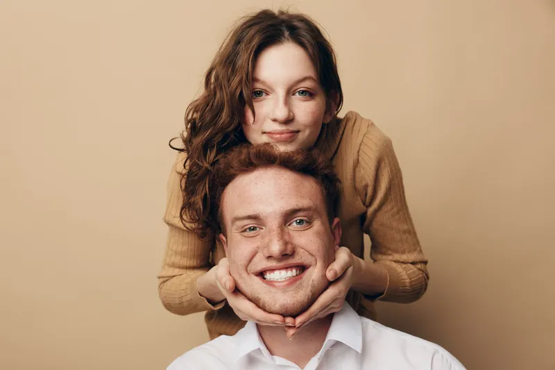 Smiling young couple with curly hair posing together on beige background. Woman stands behind man holding his face with hands. Concept of love, friendship, happiness, and relationship.