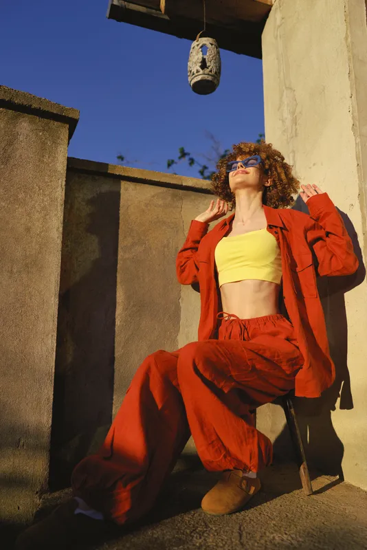 Vibrant young woman with curly hair wears a bold red suit and sunny yellow crop top, posing confidently beside a sunlit wall with a hanging lamp, radiating retro street fashion and confidence