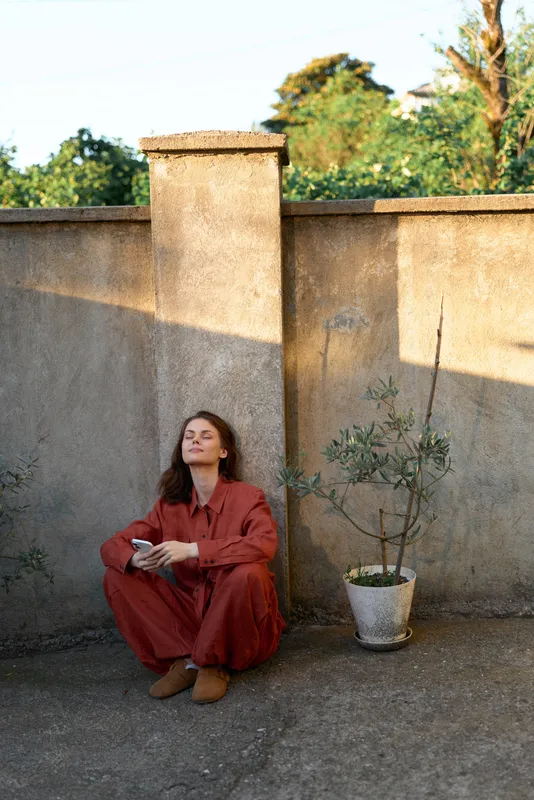 Calm woman outdoors in a red jumpsuit sits on a concrete terrace, eyes closed, holding a cup, warm light and gentle breeze enhance the relaxed lifestyle moment