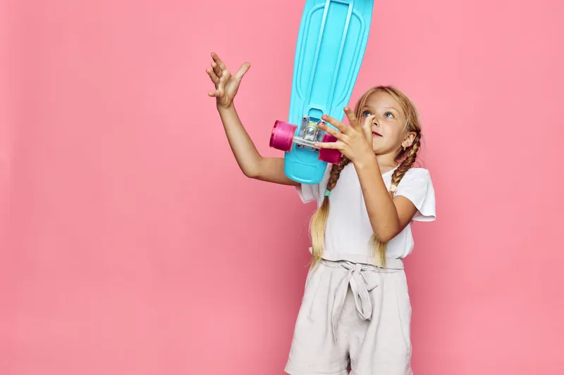 Young girl in white shirt and beige shorts balancing a blue skateboard with pink wheels on her hand. Studio shot on pink background. Concept of childhood and play.
