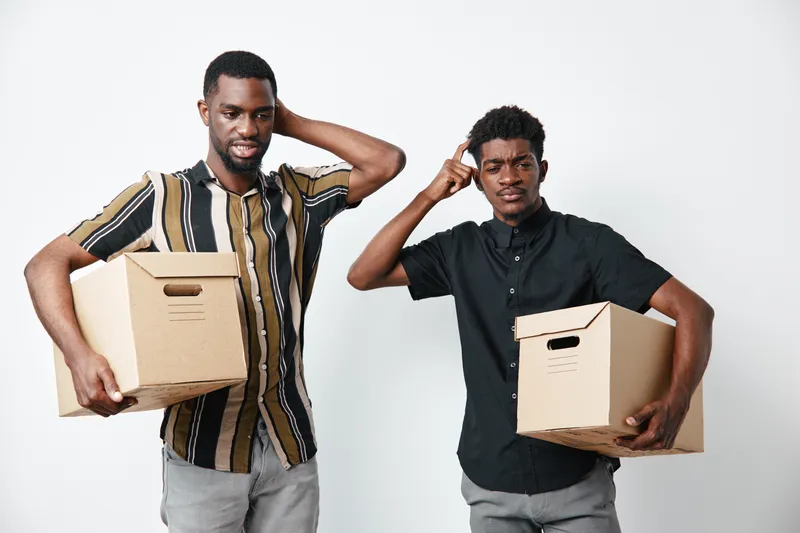 Two young black men holding cardboard boxes with confused expressions on white background. African ethnicity, dark skin, casual clothing, moving or delivery concept.