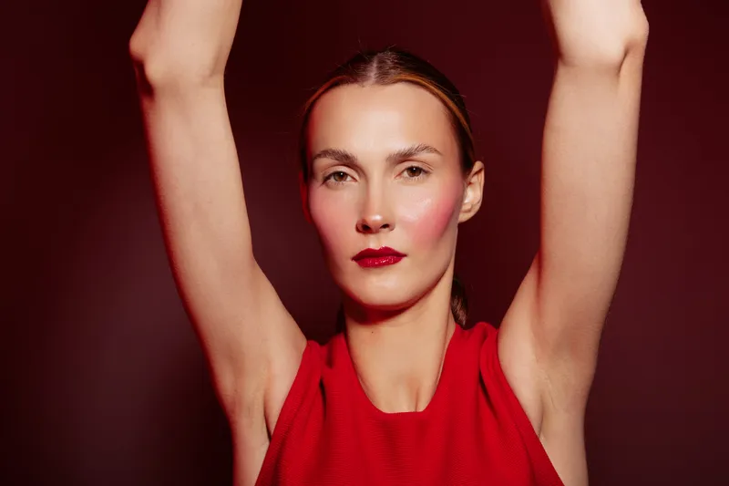 Portrait of confident woman in red sleeveless top with arms raised against dark red background. Concept of fashion, beauty, elegance, and strong female presence in studio lighting.