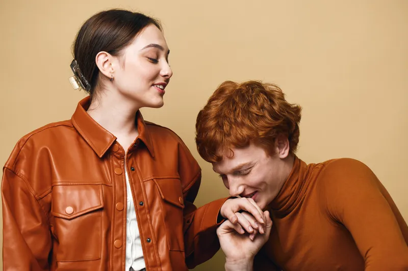 Couple in love with man kissing woman hand in romantic gesture, smiling woman wearing leather jacket and man in brown turtleneck against beige background.