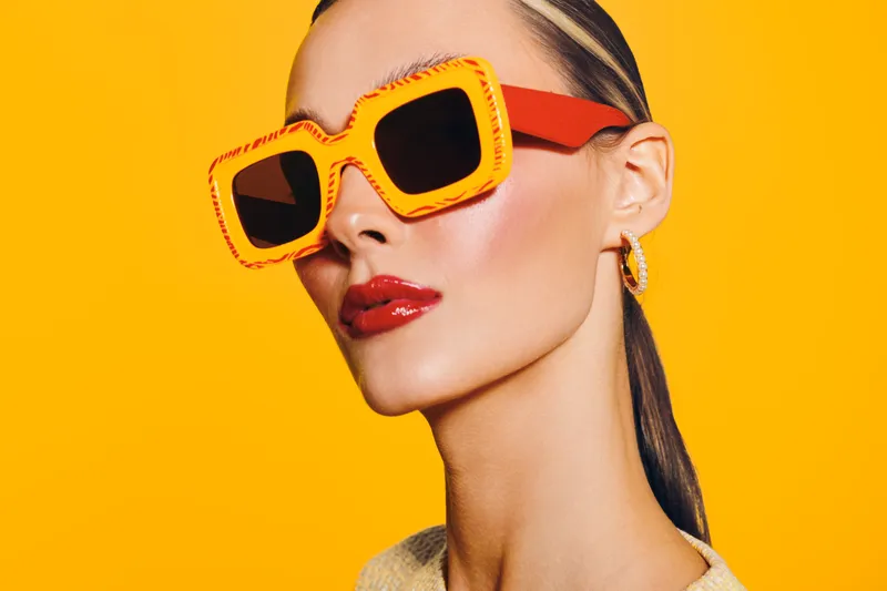 Stylish woman with slicked hair wearing oversized orange sunglasses and red lipstick poses against bright yellow background. Concept of fashion, beauty, and summer accessories.